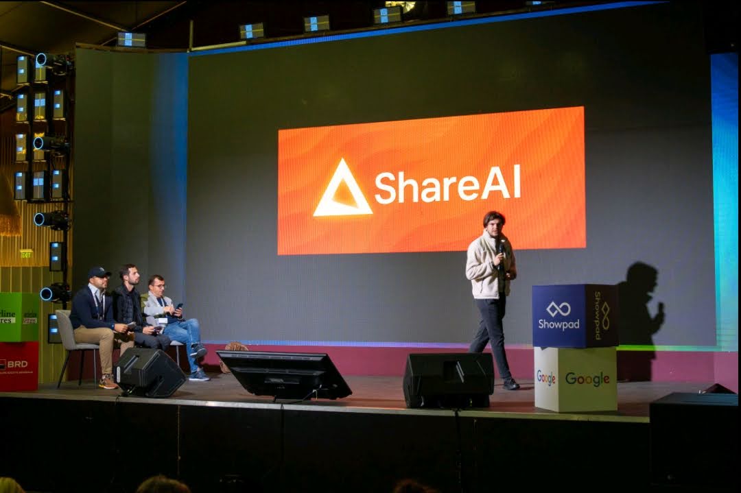 Denis Sandu presenting on stage at HTW 2025, with ShareAI on the screen behind him